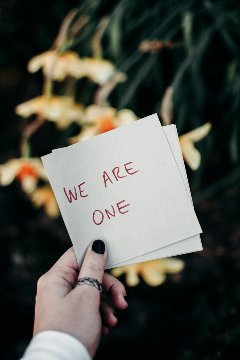 A hand holding a note reading 'We Are One' against a floral outdoor backdrop.